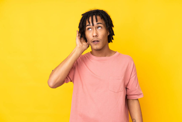 Νεαρός Αφροαμερικάνος απομονωμένος σε κίτρινο φόντο ακούγοντας κάτι βάζοντας το χέρι του στο αυτί. - Φωτογραφία, εικόνα