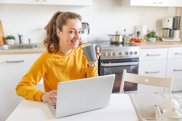 Mobilne biuro w domu. Młoda kobieta siedząca w kuchni w domu, pracująca na laptopie. Styl życia dziewczyna studiuje lub pracuje w pomieszczeniach. Koncepcja kwarantanny prowadzenia działalności na własny rachunek - Zdjęcie, obraz