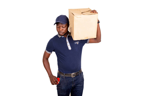 schöner junger Zusteller in blauer Uniform, der einen Karton hält und das Telefon in die Tasche steckt - Foto, Bild