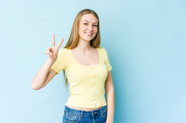 jonge blonde vrouw geïsoleerd op blauwe achtergrond vreugdevol en zorgeloos tonen een vrede symbool met vingers. - Foto, afbeelding