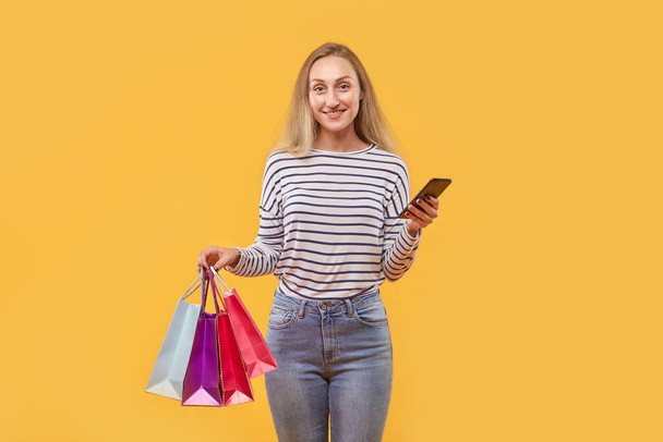 A woman looks at the camera with purchases making purchases online in her phone. - Foto, Imagem