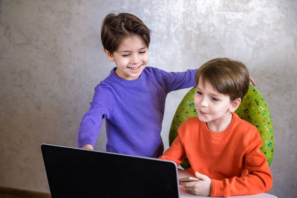 Glücklicher überglücklicher Junge mit seinem Freund, der aufgeregt schreit, während er Videospiele auf dem Laptop spielt, jubelt, nachdem er gewonnen hat, sein kleiner Bruder lächelt freudig im Hintergrund - Foto, Bild