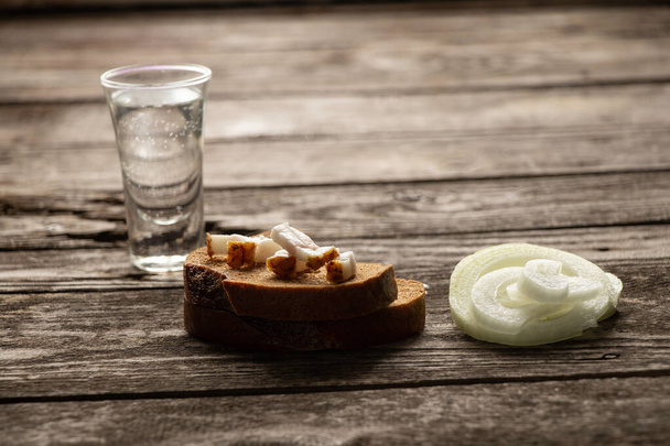 sklenice vodky vedle kusu černého chleba se slaninou na dřevěném stole v kuchyni k obědu v ukrajinštině - Fotografie, Obrázek
