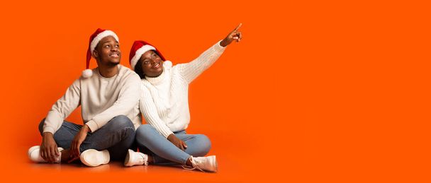 Ferienangebot. Interesantes schwarzes Paar in Weihnachtsmützen zeigt auf Kopierraum - Foto, Bild