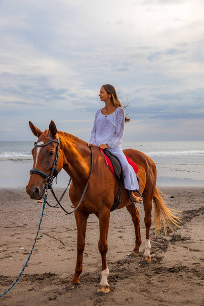 Bella donna caucasica a cavallo sulla spiaggia. Donna vestita di bianco. Ricevuto. Ora del tramonto sulla spiaggia. Attività all'aperto. Vacanze in Asia. Concetto di viaggio. Bali, Indonesia - Foto, immagini