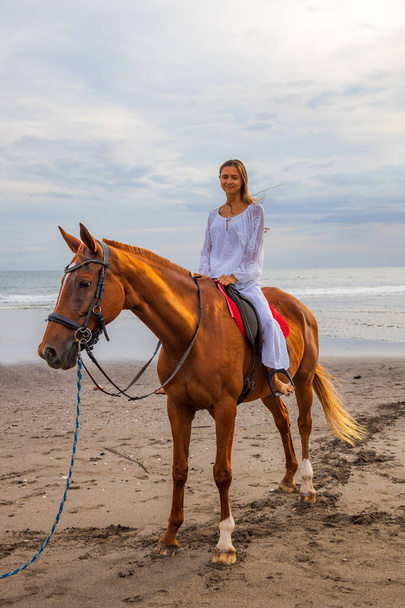 Beautiful caucasian woman riding horse on the beach. Female wearing white clothes. Copy space. Sunset time on the beach. Outdoor activities. Vacation in Asia. Traveling concept. Bali, Indonesia - Photo, Image