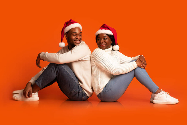 Романтическая чёрная пара в шляпах Санта-Клауса, сидящая и поворачивающаяся друг к другу - Фото, изображение