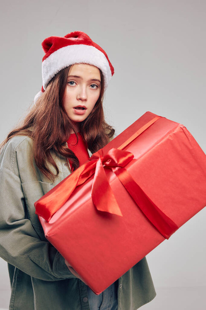 όμορφο κοριτσάκι σε ένα εορταστικό καπέλο με ένα κουτί δώρου και ένα μεγάλο χριστουγεννιάτικο φιόγκο - Φωτογραφία, εικόνα