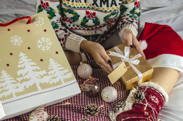 A view of a woman in a Christmas sweater holding a box with a glittery cover in her hands on the bed - Zdjęcie, obraz