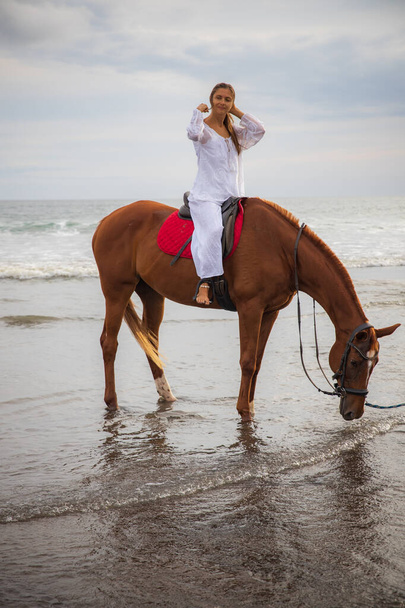 Beautiful caucasian woman riding horse by the sea. Female wearing white clothes. Copy space. Sunset time on the beach. Outdoor activities. Vacation in Asia. Traveling concept. Bali, Indonesia - Photo, Image