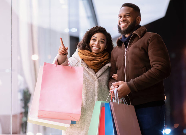 Feliz pareja negra con bolsas de compras apuntando a la ventana del centro comercial - Foto, imagen