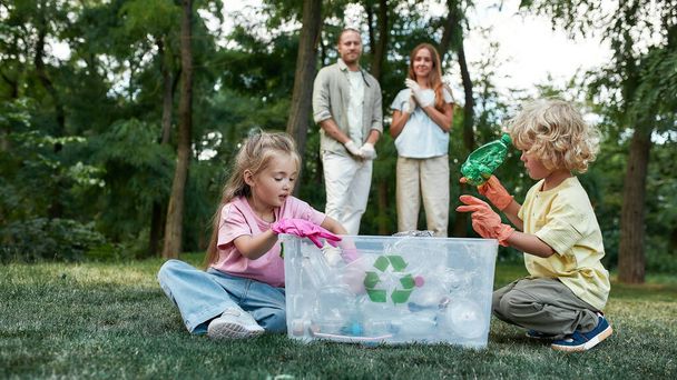 Семья Эко. Маленькие брат и сестра бросают использованные пластиковые бутылки в мусорное ведро во время сбора пластиковых отходов в лесу или парке с родителями - Фото, изображение