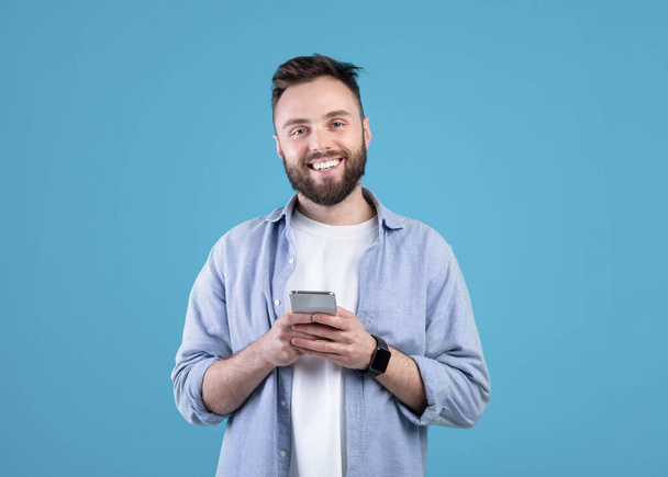 Portré hűvös szakállas fickó használ mobiltelefon kék stúdió háttér - Fotó, kép