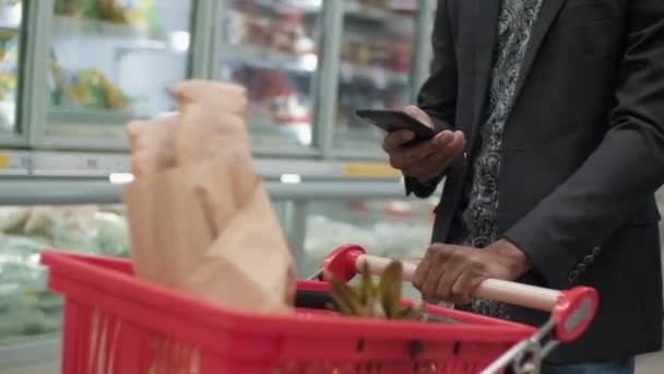 Midsection footage of unrecognizable man in suit using smartphone while carrying shopping cart with products walking along hypermarket - Footage, Video