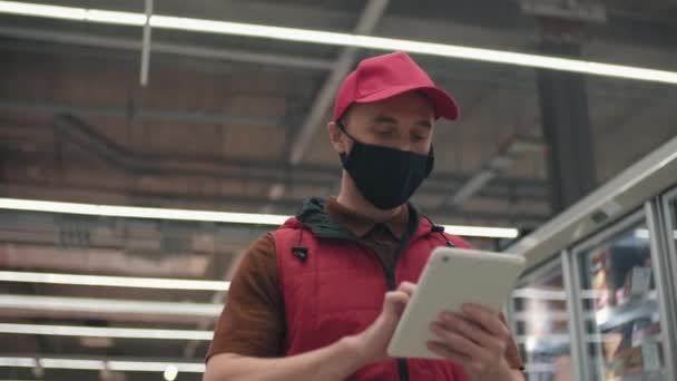 Low-angle slow-mo medium shot di merchandizer di ipermercato in maschera uniforme e protettiva passeggiando lungo i corridoi con tablet analizzando le vendite di magazzino - Filmati, video