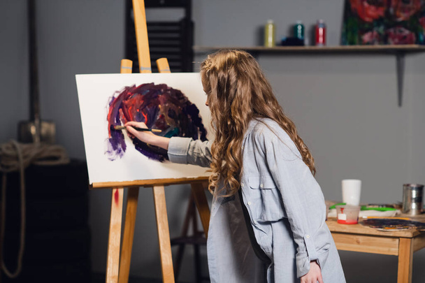 Ένα νεαρό χαριτωμένο κορίτσι καλλιτέχνης μαθαίνει να ζωγραφίζει σε ένα εργαστήριο τέχνης. Σχεδιάζει ένα καβαλέτο με λαδομπογιές και ένα πινέλο. - Φωτογραφία, εικόνα