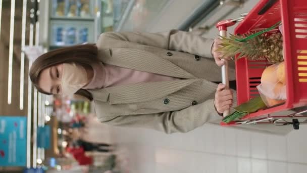 Vertikale mittlere Aufnahme einer smarten, geschäftstüchtigen asiatischen Frau in Maske, die einen Einkaufswagen mit Produkten trägt, die durch die Gänge eines großen Supermarktes gehen und täglich Lebensmittel einkaufen - Filmmaterial, Video