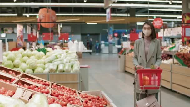 Korumalı maske takmış iş adamı bir kadının, karantinada yiyecek alışverişi yaparken taze domates toplarken, büyük süpermarketin market bölümünde el arabasıyla eko-torbaya koyduğu orta boy bir fotoğraf. - Video, Çekim