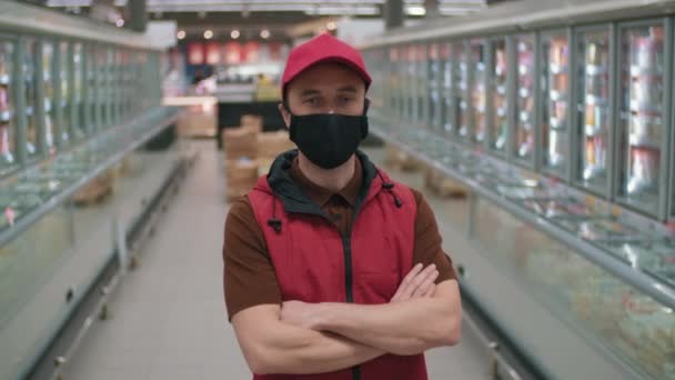 Plan moyen d'homme caucasien debout avec les mains pliées portant un uniforme rouge et un masque de protection travaillant comme marchand dans un supermarché regardant une caméra avec des boîtes déballées avec des marchandises en arrière-plan - Séquence, vidéo