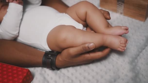 Χαριτωμένο μωρό πόδια στα χέρια του μαύρου πατέρα που περιβάλλεται από χριστουγεννιάτικα δώρα, κοντά - Πλάνα, βίντεο