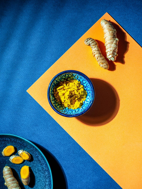 Turmeric (curcumin, Curcuma Longa Linn), powder in blue ceramic bowl and rhizome on orange and blue background. Overhead shot with copy space. - Photo, Image