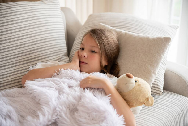 Una bambina malata e preoccupata coperta di coperte sta abbracciando un orsacchiotto.. - Foto, immagini
