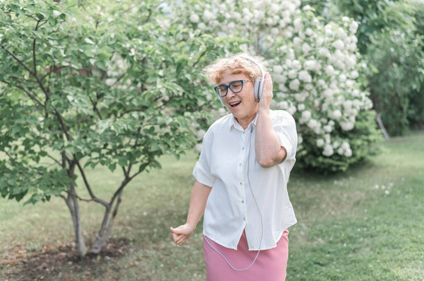 Happy old woman listens to music on headphones and dances in a park of blooming apple trees. Active female pensioner smiles and enjoys a warm summer day in the garden. Advanced older generation. - Foto, Bild