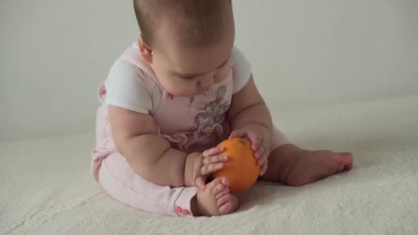 младенец, детство, питание, первое кормление, витамины концепция - радость веселая игривая ребенок 8 месяцев девочка ест оранжевый. пухлый ребенок берет круглые сладкие вкусные фрукты с руками в помещении на белом фоне - Кадры, видео