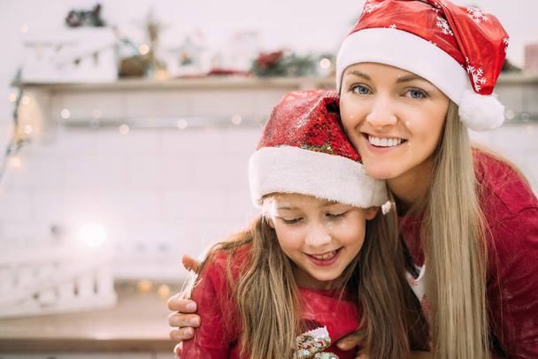 Junge Mutter und Teenager-Tochter in der Küche mit roter Nikolausmütze lachen und ihre Gesichter sind mit Mehl bedeckt. Weihnachtliches Kochkonzept. - Foto, Bild