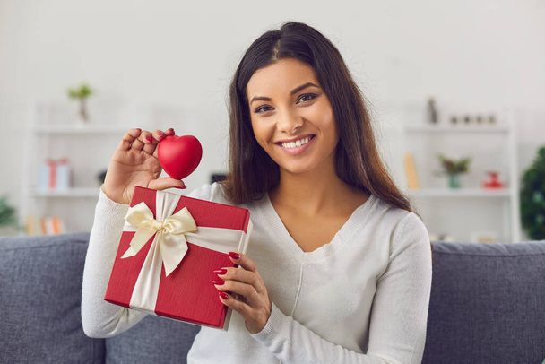Happy smiling pretty brunette girl showing holiday present box with ribbon and red heart - Фото, зображення