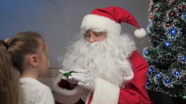 Joulupukki istuu sohvalla pienen tytön kanssa, joulupukki antoi lahjan leluautolle, lapsi iloitsee ja halailee maagista isoisää. loma ja juhla. Perheiden lapset talviloma. Hyvää jouluaattoa.. - Materiaali, video