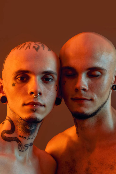 Close up portrait of young half naked twin brothers with tattoos and piercings posing together isolated over orange background - Foto, Imagem