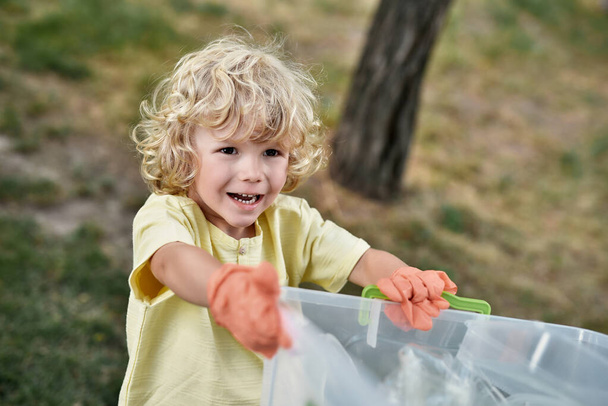 Cute little boy wearing rubber gloves holding recycle bin and smiling while collecting plastic waste in forest or park with parents - Photo, image