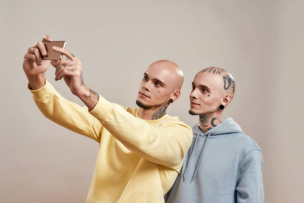 Photo with best friend. Two young caucasian twin brothers in casual wear taking selfie on mobile phone while standing together isolated over beige background - Foto, Imagem