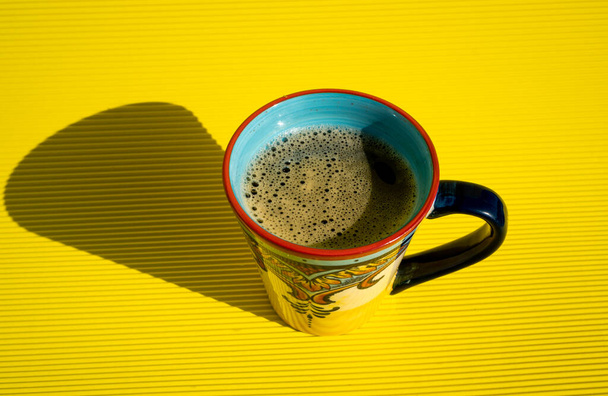 Exquise vers gemaakte mousserende koffie in een blauwe handgemaakte beker weerspiegelt een zwarte schaduw aan de zijkant op een gele tafel. - Foto, afbeelding