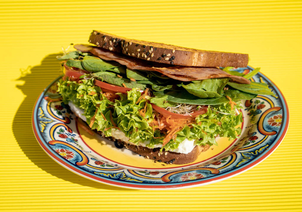 Delicious healthy ham sandwich with whole wheat bread, tomato, lettuce, avocado and cheese inside on a handmade plate on a yellow table. - Foto, afbeelding