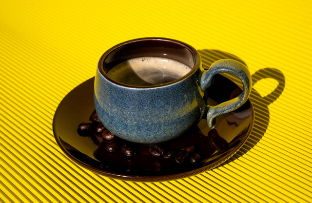 黄色のテーブルの下にコーヒー豆と茶色のプレートの上に青のカップでおいしい、新しく醸造されたアメリカのコーヒー. - 写真・画像