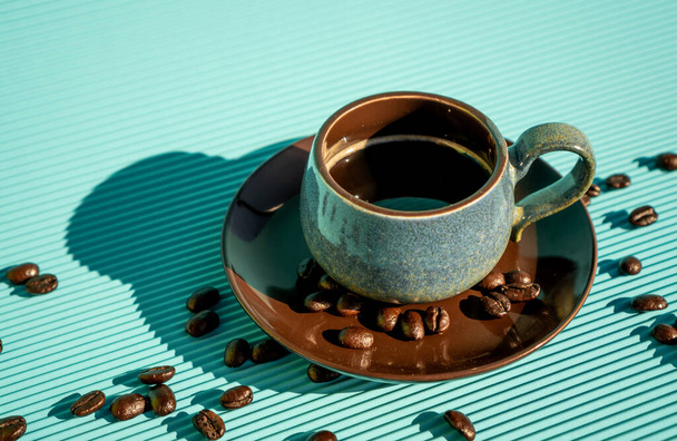 Köstlicher, frisch gebrühter amerikanischer Kaffee in einer blauen Tasse auf einem braunen Teller, darunter Kaffeebohnen auf einem blauen Tisch. - Foto, Bild