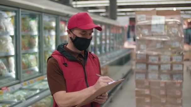 Gros plan moyen de l'homme en uniforme rouge travaillant comme marchand dans un grand hypermarché écrivant quelque chose sur le presse-papiers - Séquence, vidéo