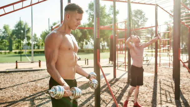 Muscular men training muscles at outdoor gym. Young guy with naked torso working out using dumbbells on a sunny day - Fotó, kép
