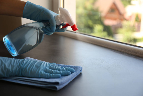 Woman in gloves cleaning grey stone window sill with rag and detergent indoors, closeup - Foto, Imagen