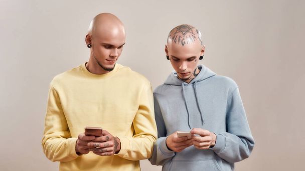 Two young caucasian twin brothers in casual wear with tattoos and piercings using smartphones while standing isolated over beige background - Photo, Image