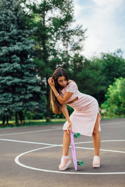 Portrait of a smiling charming brunette female holding in hand her skateboard on a basketball court. Happy woman with trendy look taking break during sunset. - Fotografie, Obrázek
