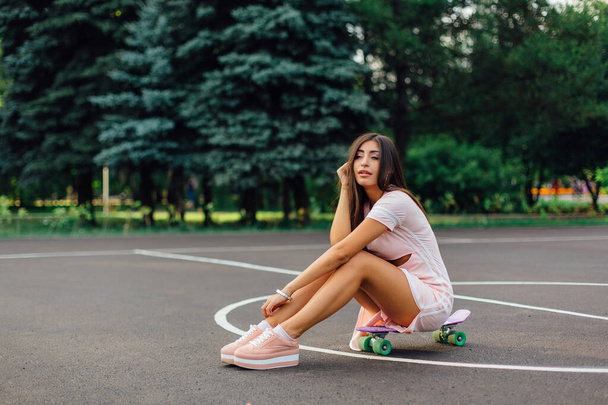 Portrait of a smiling charming brunette female sitting on her skateboard on a basketball court. Happy woman with trendy look taking break during sunset. - Valokuva, kuva