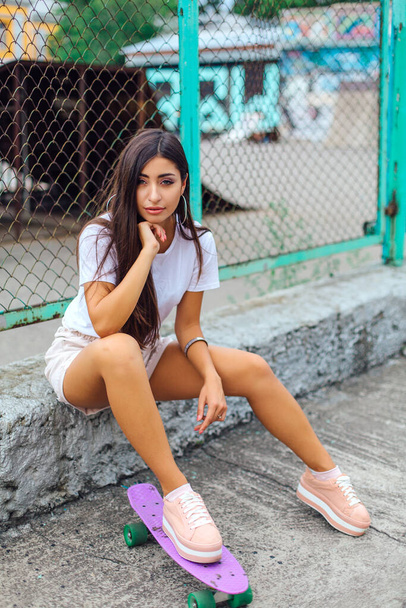 Summer lifestyle image of trendy pretty young girl sitting next to the skateboard coart with her purple plastic skateboard. - 写真・画像
