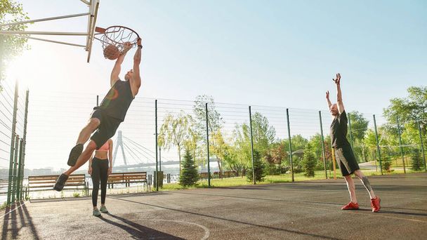 Full length shot of active young people spending time outdoors on playground. Skilled sportive guy throwing ball into basket, while playing basketball - Photo, Image
