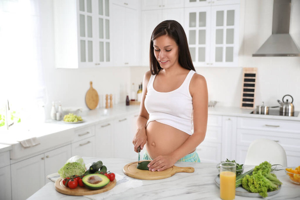 Young pregnant woman cutting avocado at table in kitchen. Taking care of baby health - Fotoğraf, Görsel