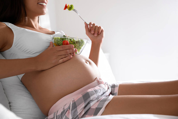 Young pregnant woman with bowl of vegetable salad in bedroom, closeup. Taking care of baby health - Foto, imagen