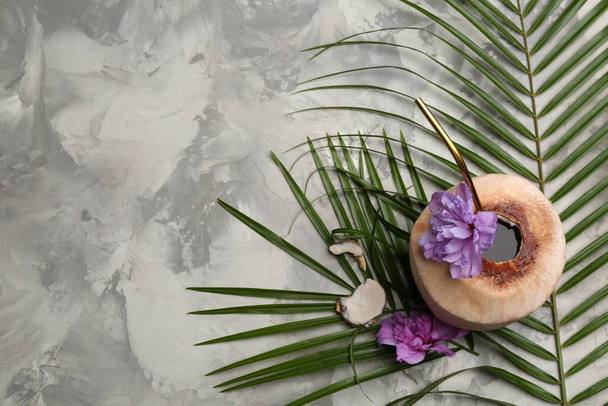 Young peeled coconut with straw, tropical leaf and flowers on grey table, flat lay. Space for text - Фото, изображение
