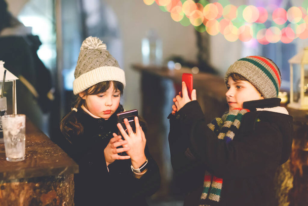 2人の小さな子供、男の子と女の子は、携帯電話や携帯電話で写真を撮るという強い雪の中で伝統的なクリスマスマーケットで楽しんでいます。幸せな子供、兄弟、親友 - 写真・画像